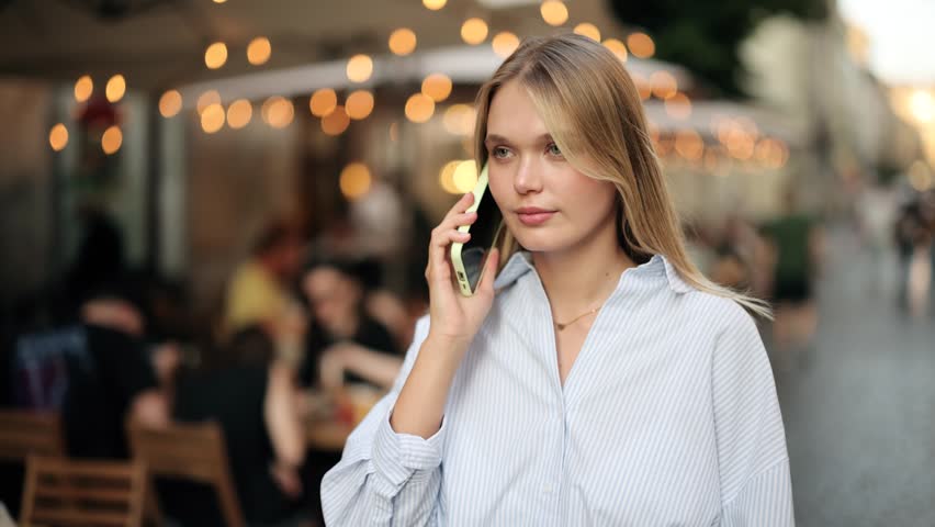 Confused blonde female pedestrian talking on smartphone with shocked facial expression, urban background