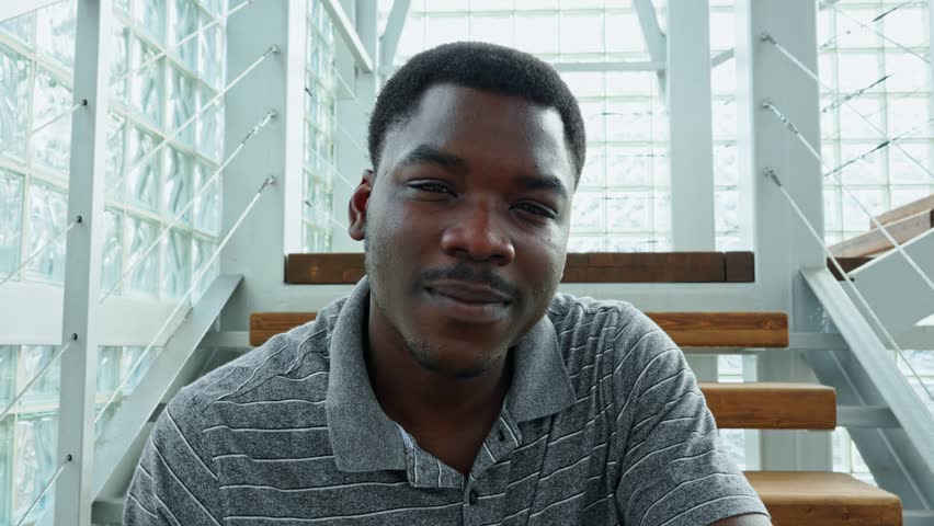 Smiling man adjusts glasses while seated inside modern building with glass walls and wooden stairs