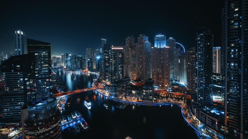 Wide angle timelapse of Dubai Marina waterfront and skyline at night with traffic flowing across bridge and boats cruising with Ain Dubai ferris wheel glowing in the distance