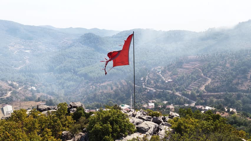 Aerial Video Torn Red Flag Over Burned Forest After Wildfire at Daytime Drone shot of a damaged red flag waving in strong wind above a mountain landscape covered with smoke after a wildfire, showing destruction and environmental impact
