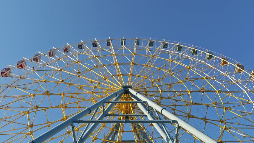 Huge ferris wheel with colorful cabins rotating at an amusement park. Low angle view of the spinning ride against a bright, cloudless sky