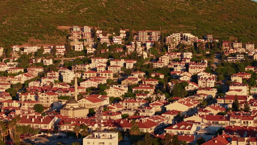 Aerial pan of residential hillside town with red terracotta roofs and mosque minaret during golden hour, marina of Datcha Turkey.