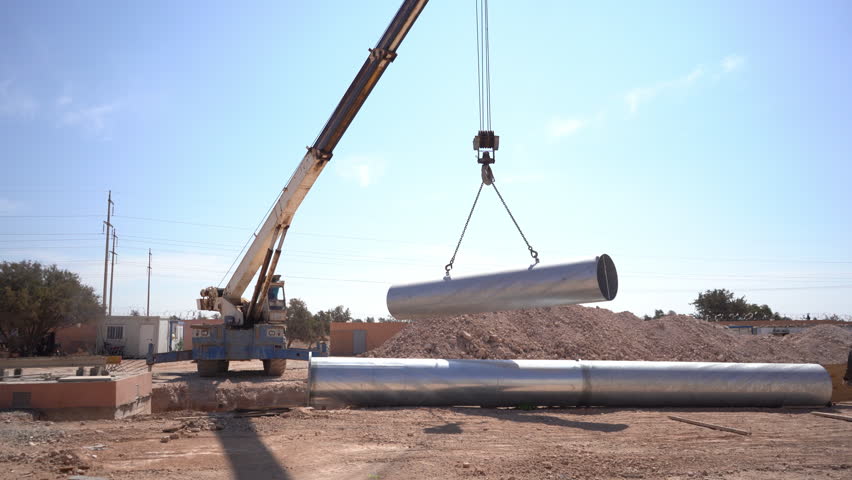 A high-quality wide shot capturing a heavy-duty mobile crane lifting a massive, reflective metal pipe at a busy industrial construction site. Suspended by strong steel cables and hooks, the large cylindrical tube is carefully maneuvered into position, likely for an underground pipeline, water purification system, or municipal drainage infrastructure project. Another identical steel pipe rests on the ground next to a freshly dug trench and large piles of excavated dirt, set against a clear, vibrant blue sky. This dynamic B-roll footage is an excellent commercial asset for projects focusing on civil engineering, industrial construction, heavy machinery operations, and public utility development. It perfectly illustrates the large-scale physical labor and specialized equipment required to build modern, sustainable infrastructure. Ideal for corporate presentations, engineering documentaries, and construction company promotional videos.