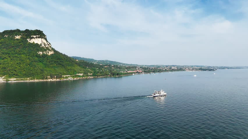 Passenger ferry moving across Lake Garda with mountain slope and shoreline in background, aerial view