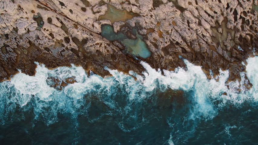Waves crashing onto a textured rocky shoreline with tide pools from above