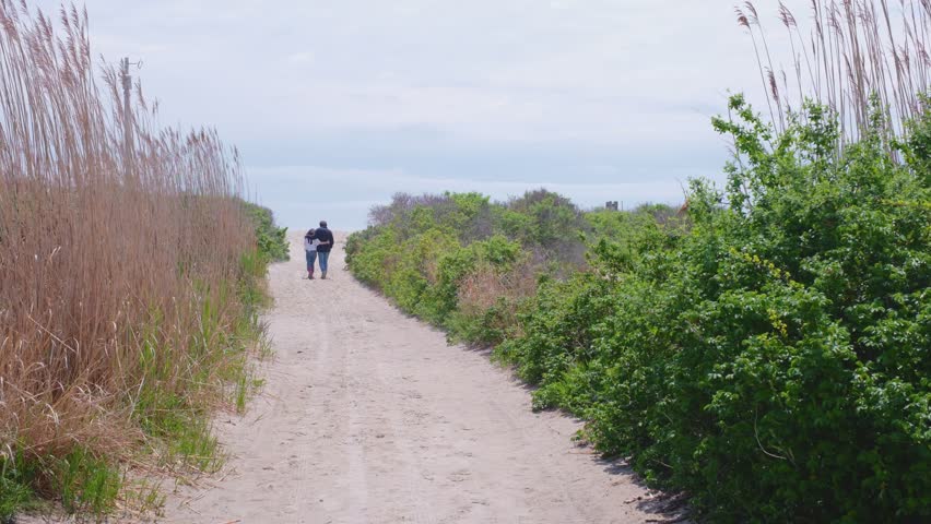 Two people walking down a sandy path toward the beach on a bright, overcast day.