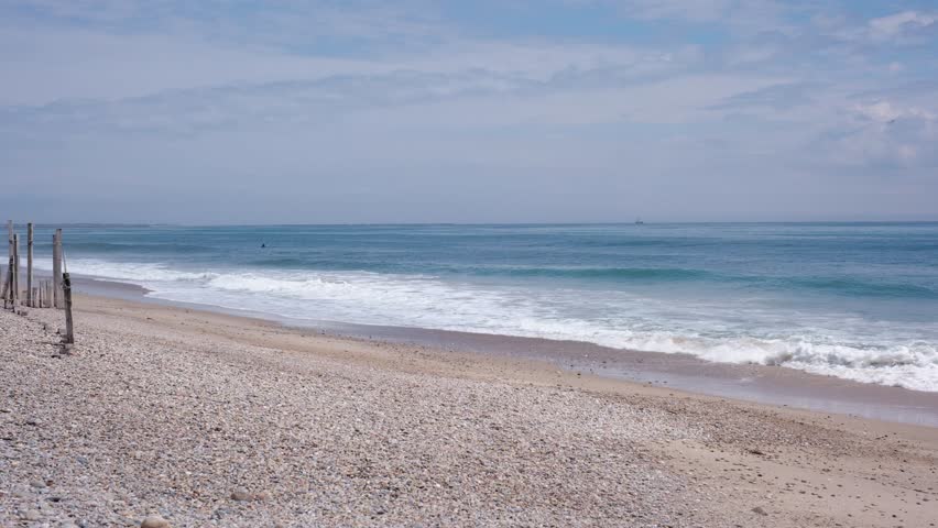 Ocean waves rolling onto a wide pebble and sand beach shoreline under a cloudy sky.