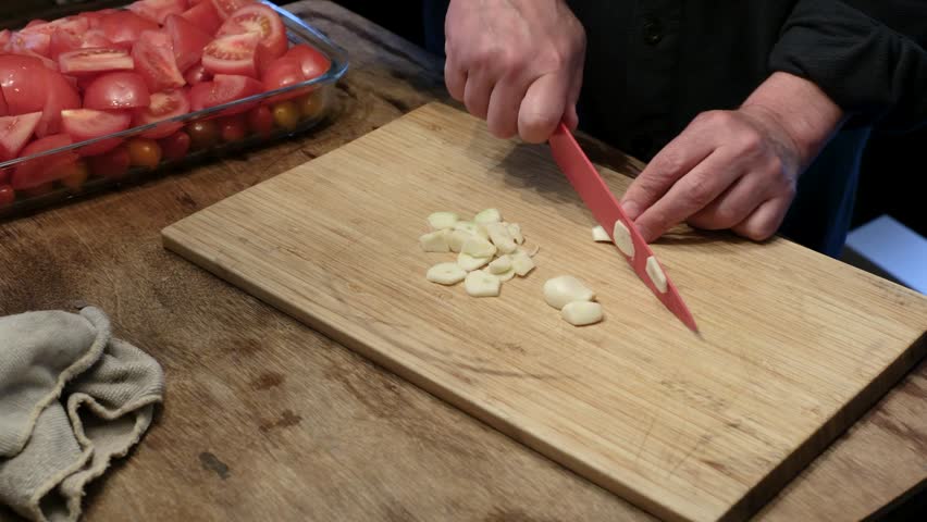 Close up of a chef slicing fresh garlic cloves with a red knife on a wooden cutting board.
