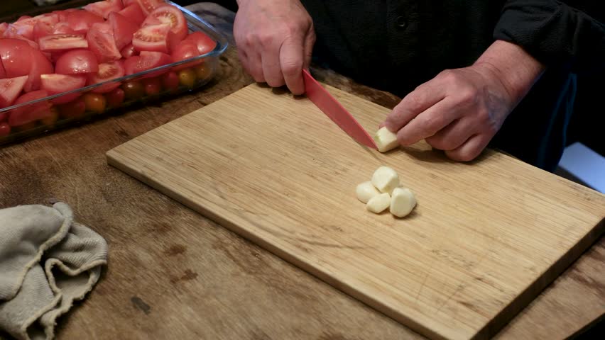 Close up of a chef slicing fresh garlic cloves with a red knife on a wooden cutting board.