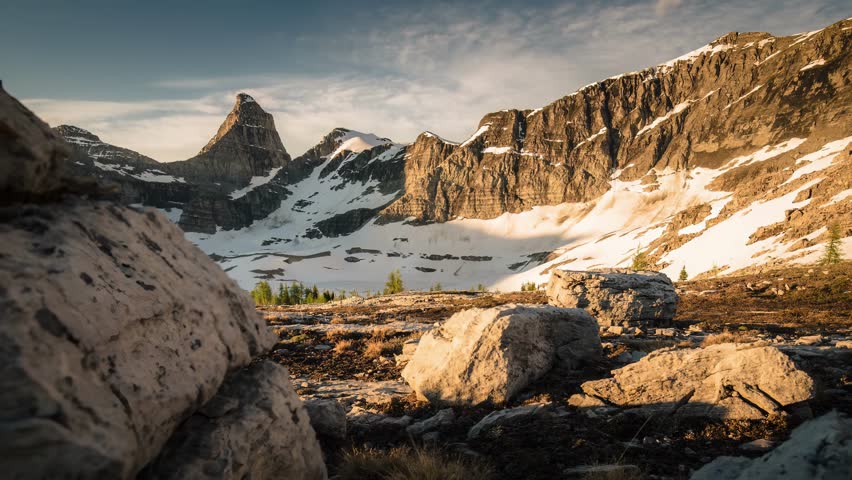 Dramatic mountain landscape at sunset, showcasing rugged terrain, patches of snow, and sparse vegetation under a vast sky