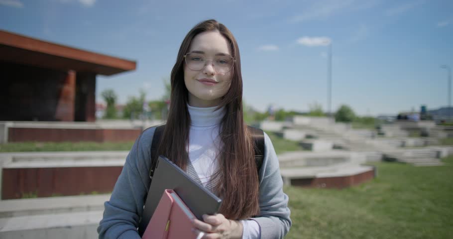 A hand-held portrait of a young female student in casual clothes, wearing glasses and a backpack, holding a notebook and laptop in her hands and making a welcoming gesture with her hands - victory