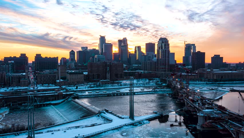 Dynamic aerial drone footage captures minneapolis downtown buildings and icy mississippi river during a breathtaking winter sunset, featuring vibrant colors in sky and cityscape.