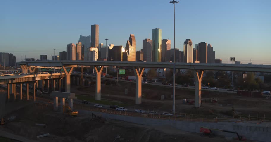 Cinematic aerial view of Houston Texas city skyscrapers glowing in golden light during sunset. Camera pulls back revealing massive highway infrastructure projects and crane work in urban area.