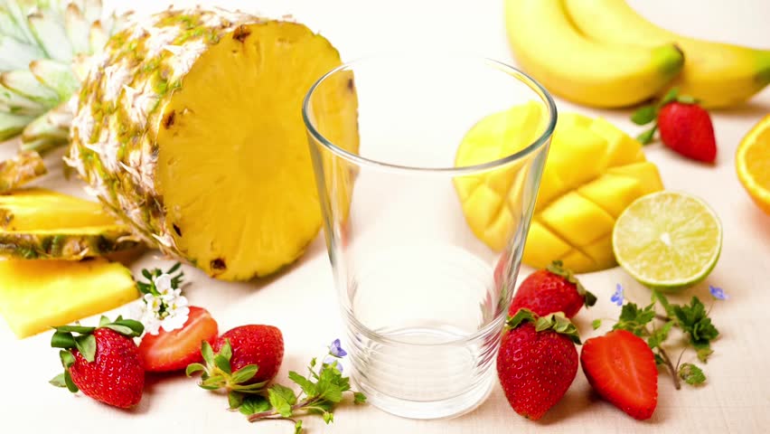 pink fruit smoothie pouring in glass with straw surrounded by fresh pineapple, mango, strawberries and bananas, healthy tropical breakfast beverage