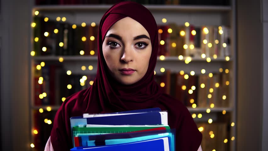 Woman in hijab holding stack of files in front of glowing lights, serious determined gaze and composed posture, studio evening portrait conveying ambition, planning, and leadership tone