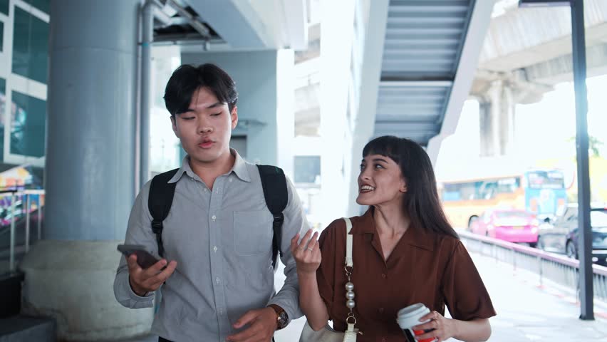 Asian man using a phone walks with a woman holding a coffee cup at a transit station. Two office worker talk during a commute on a city sidewalk