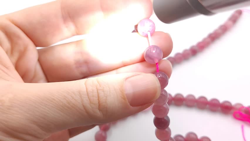 A person meticulously inspects translucent pink quartz crystal beads on a string under a bright examination lamp to verify quality and clarity for jewelry making supplies and custom designs