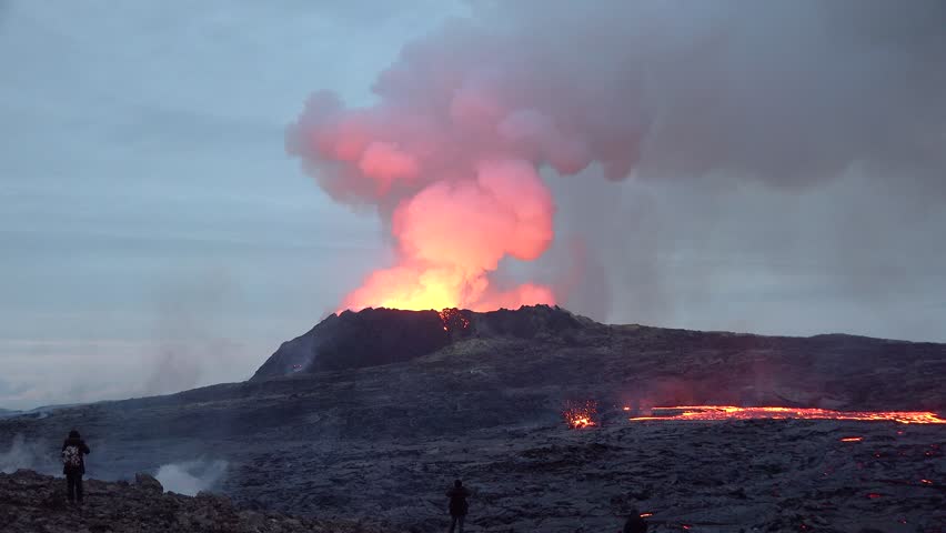 A volcano erupts at Fagradalsfjall in Iceland, sending lava and smoke into the sky. People look on as the volcano continues to flow lava down the sides in the evening light.