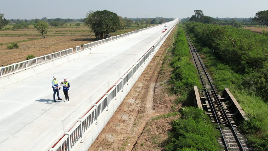 Long concrete bridge over rural field with railway track and roadway construction, engineer workers on site, aerial view of new bridge in landscape under construction