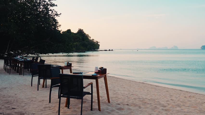 Restaurant tables and chairs arranged on sandy beach, tropical coast during golden hour sunset, calm ocean waves, luxury travel background, romantic dining setup