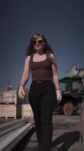 Young woman wearing sunglasses and work gloves standing near agricultural machinery outdoors on sunny day. Modern farming lifestyle and agriculture industry concept with rural background.