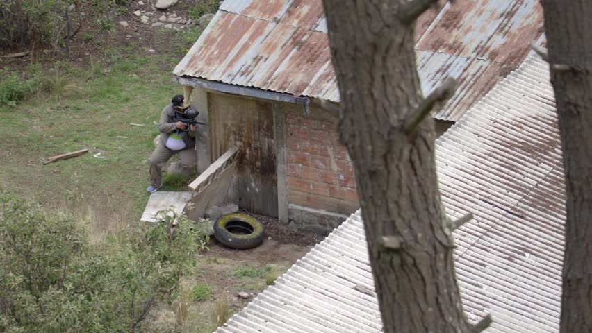 In paintball, behind a brick house, a man hides while watching with his gun, protective helmet, and green uniform. The house is rustic, brick-colored with a corrugated iron roof, in a natural and wild space.