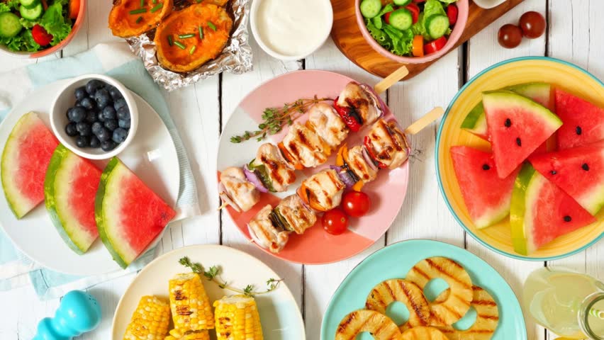 Summer BBQ or picnic food table scene over a white wood background. Slow rotating zoom motion. Selection of fruits, salad, grilled meat and potatoes.