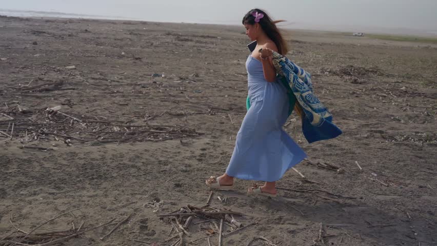 Beautiful young woman wearing a blue dress smiling and walking on a deserted sandy beach. The wind is blowing her hair and her blanket