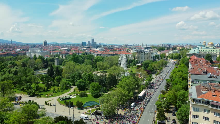 Aerial drone footage of a large group of cyclists riding through central Sofia, Bulgaria, along a major city boulevard on a sunny spring day. Wide urban skyline view featuring city park, Ferris wheel, blue sky, and active outdoor recreation.