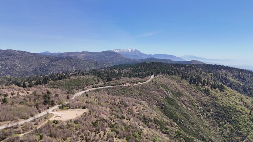 A wide drone shot of a sweeping curve on the Rim of the World Highway, with snow-dusted San Bernardino peaks visible in the far distance.