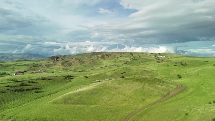 Stunning aerial view of expansive green hills and mountains under a dynamic cloudy sky, with distant wind turbines generating clean, renewable energy in a serene and natural environment