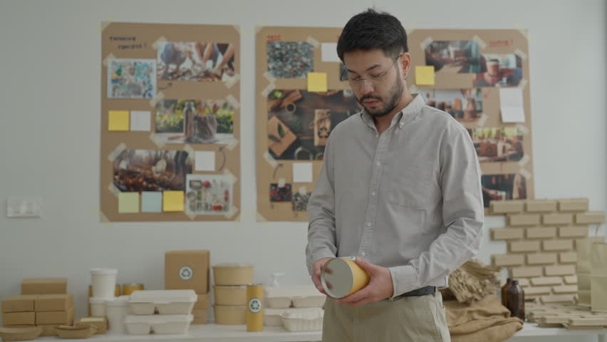 Man holding yellow cup office workspace with bulletin board, project notes, desk, shirt, and businessman holding paper tape with packaging samples, planning board, cardboard tube, thoughtful