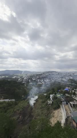 High angle panoramic view of a valley town nestled among hills under a dramatic, overcast stormy sky. Plumes of white smoke rise from a hillside bonfire in the foreground, with agricultural fields and residential houses stretching across the distant landscape.