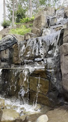 Close-up view of a multi-tiered artificial waterfall cascading down dark, rough stone boulders. Water flows over the rocky steps in a landscaped garden with trees and green plants visible at the top.
