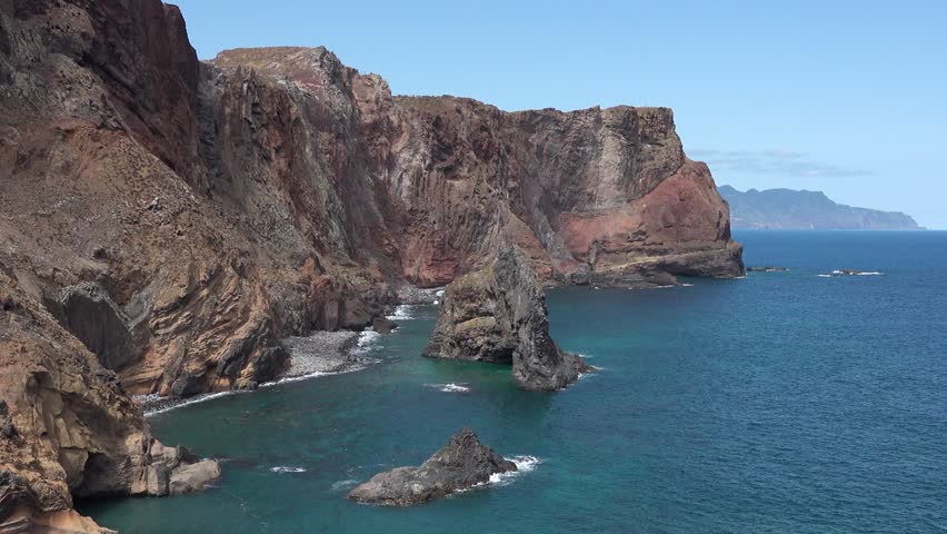 Cliffs rise sharply from the ocean at Madeira island. The waves crash against the rocks below as the sun shines overhead.
