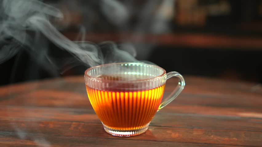 Hot amber tea in transparent glass cup with steam, cozy warm light, dark wooden table, soft bokeh background, slow motion