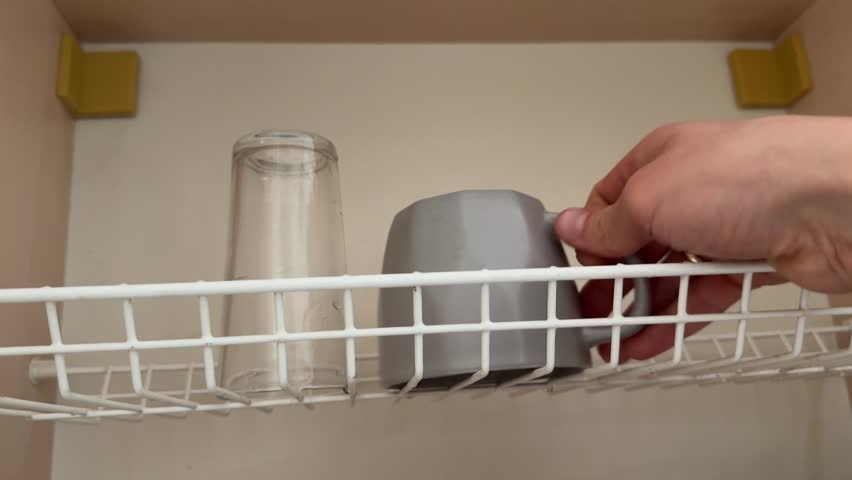 A person places a gray ceramic mug onto a metal wire shelf inside a kitchen cabinet.