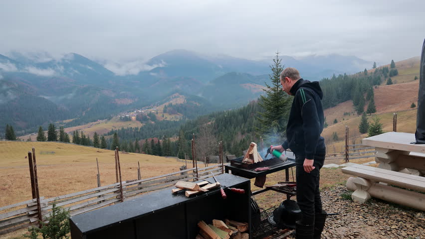 Man Grilling Meat In Mountain Valley Landscape, outdoor barbecue video frame with smoke, wooden table and misty hills behind