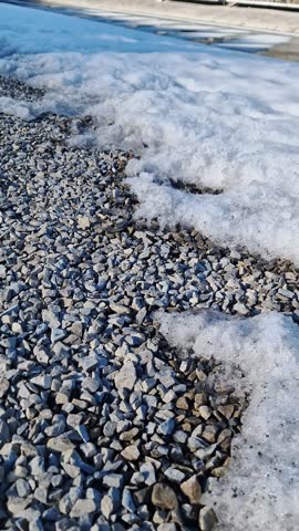Camera tilt reveals melting snow on gravel leading to a mountain lake with a Ferris wheel and resort in Bukovel. Concept of seasonal transition, mountain tourism, and spring awakening.