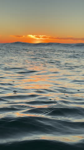 A warm orange sun rises over a dark rippled ocean surface. Captured in slow motion within the sea as waves roll towards the beach.
