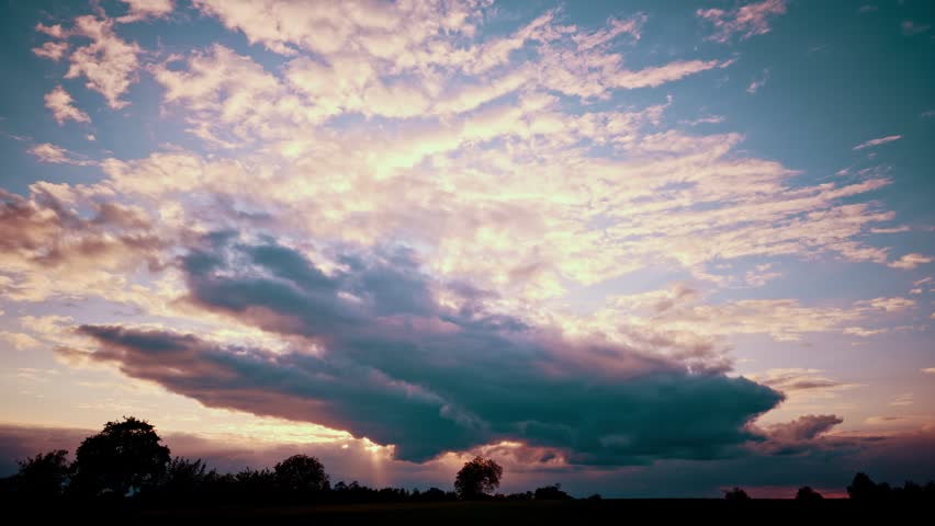 Dramatic sunset sky with colorful clouds in pink, purple, and blue, sun rays breaking through dark clouds above a silhouetted tree line.