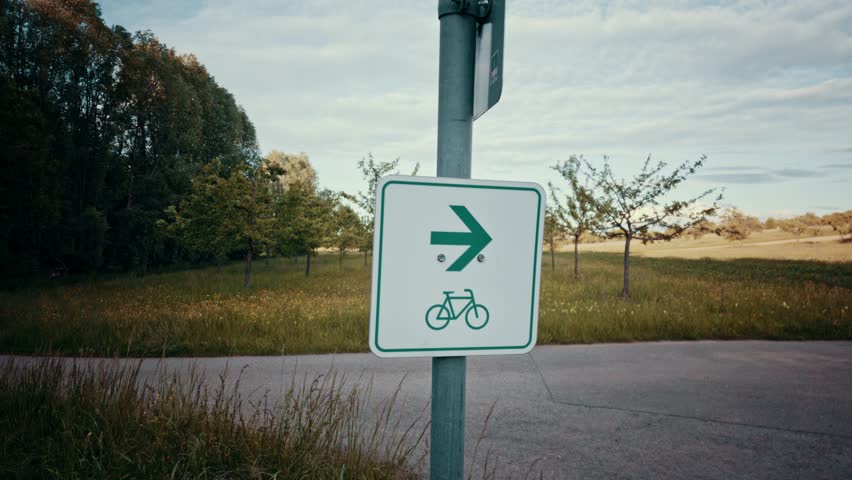 Sign with green arrow and bicycle icon marking a cycle path to the right, leading through a grassy field with trees under a cloudy sky.