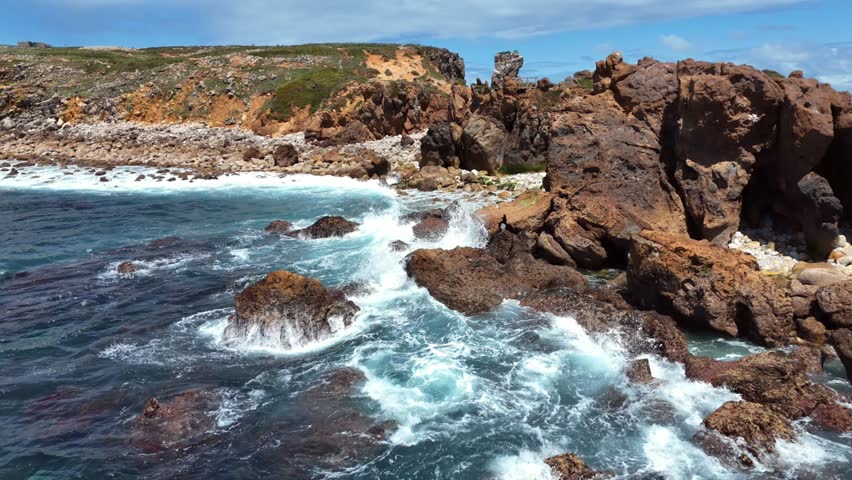 Low Angle View of Turquoise Waves Washing Over Mossy Boulders Below Eroded Cliff With Wooden Viewpoint