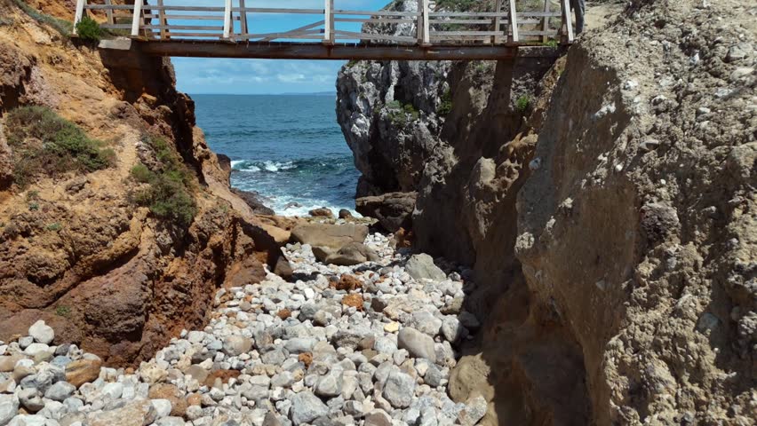Wooden Footbridge Spanning Rocky Coastal Ravine With Pebble Beach and Ocean View Below