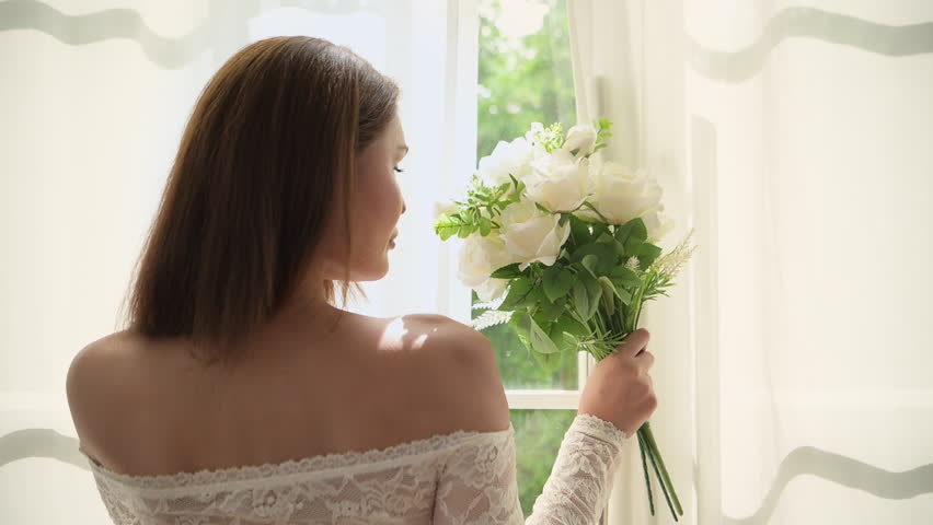 Beautiful bride in an elegant wedding dress holding a bouquet by the window with warm sunlight shining through, preparing for her wedding ceremony in a romantic and joyful atmosphere