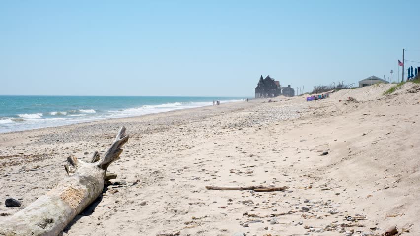 A large piece of driftwood rests on a sandy beach in Rhode Island with a historic building in the distance.