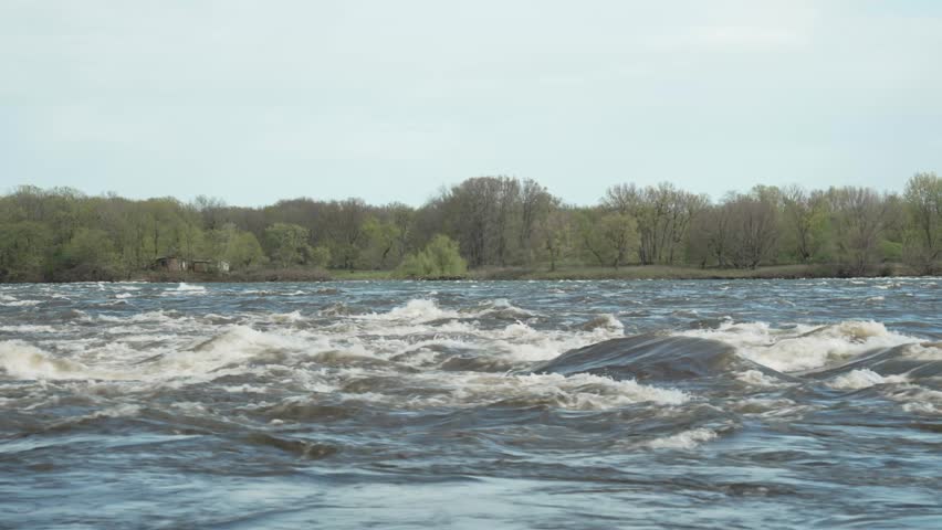 A fast-flowing river in Rapid Park with strong currents and white foaming water rushing downstream. Across the river, a low forested area creates a calm contrast to the turbulent flow. The scene captures natural outdoor ambience with dynamic water movement and peaceful green landscape in the background.
