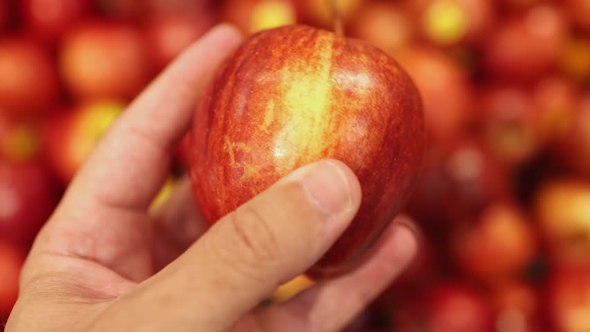 Hand holding a fresh ripe red apple fruit in a supermarket. Close-up of organic sweet agriculture product on a blurred grocery store display background.