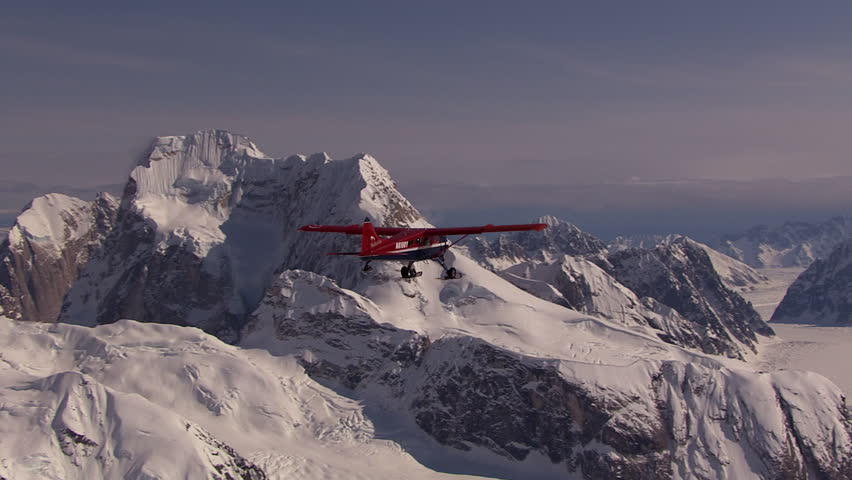 Red single propeller airplane flying above a vast, snow covered mountain range and glacier