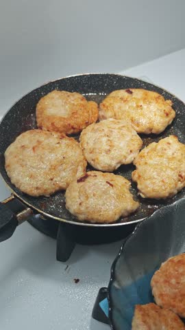 Close-up vertical shot of delicious homemade ground meat or fish patties frying and sizzling in hot oil in a black non-stick pan on a gas stovetop, gradually turning golden brown, highlighting the